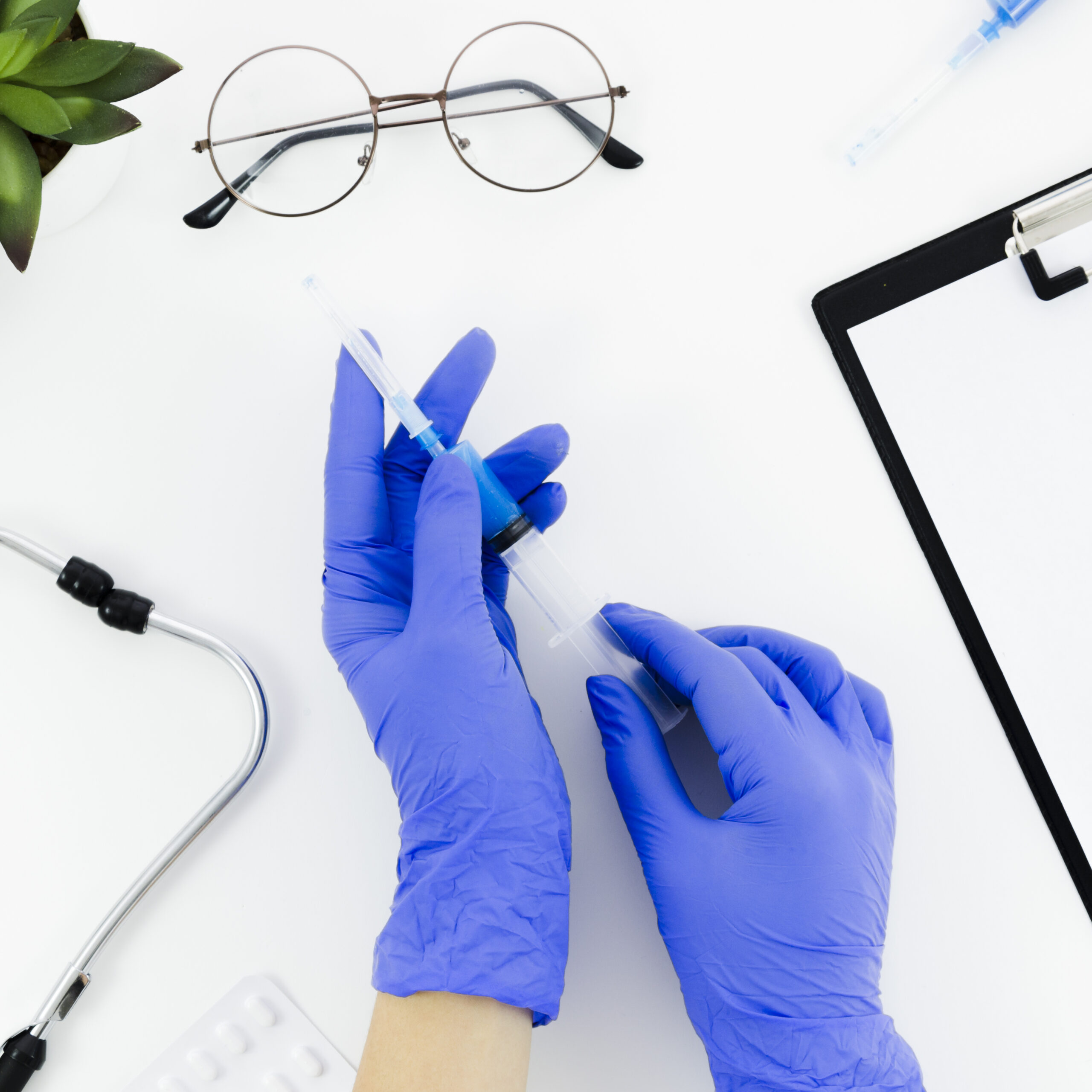 doctor s hand wearing blue gloves holding syringe white desk with stethoscope eyeglasses pills clipboard scaled